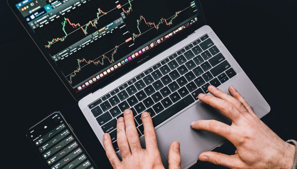 Hands analyzing stock charts on a laptop with a smartphone displaying financial data