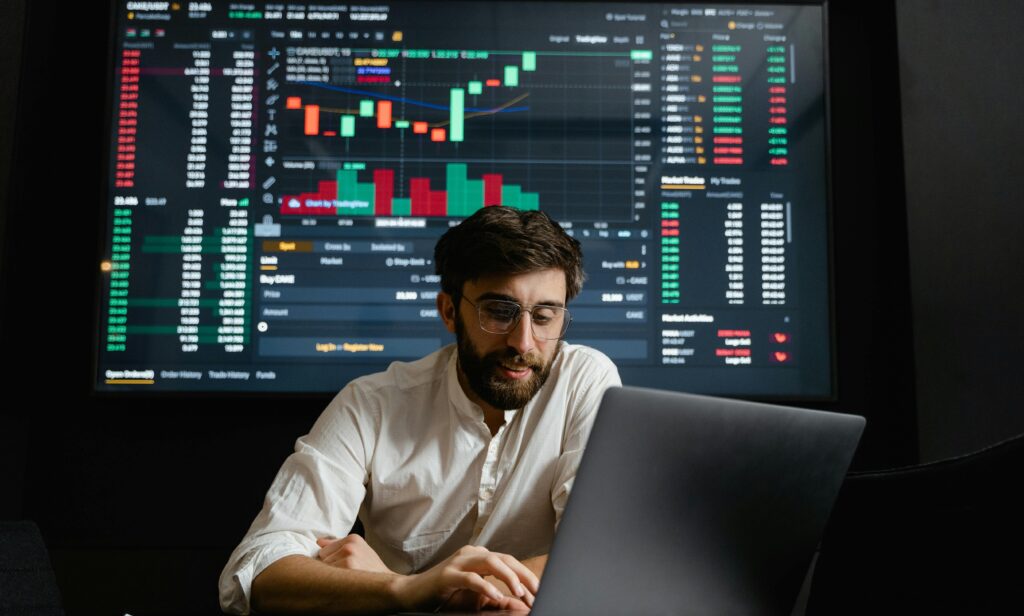 Stock trader analyzing financial charts on a laptop with market data on a screen