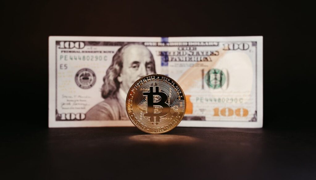 Bitcoin coin standing in front of a $100 bill, representing the relationship between traditional currency and cryptocurrency.